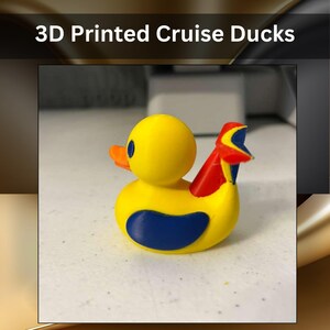 May include: A 3D printed yellow duck figurine with a blue wing, an orange beak, and a red and blue tail attachment. The text 3D Printed Cruise Ducks is displayed above the object, which features a smooth, matte finish and rounded shape.
