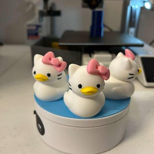 May include: Three white duck-shaped figurines with cat-like ears, black eyes, yellow beaks, and pink bows on their heads. The figurines are arranged on a circular white base with a blue top surface, positioned in front of a 3D printer.