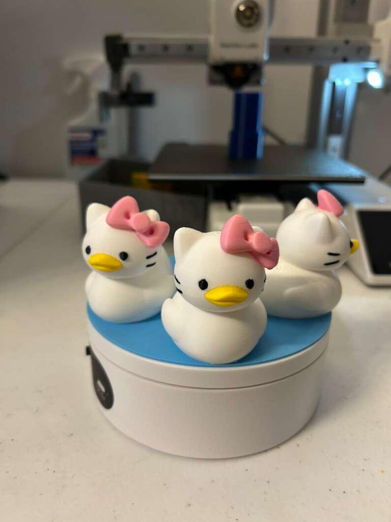 May include: Three white duck-shaped figurines with cat-like ears, black eyes, yellow beaks, and pink bows on their heads. The figurines are arranged on a circular white base with a blue top surface, positioned in front of a 3D printer.