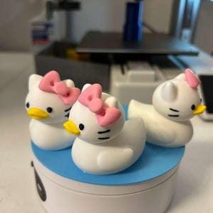 May include: Three white duck-shaped figurines with cat-like ears, black eyes, yellow beaks, and pink bows on their heads. The figurines are arranged on a circular blue and white platform, displaying a hybrid design of a duck and a cat character.