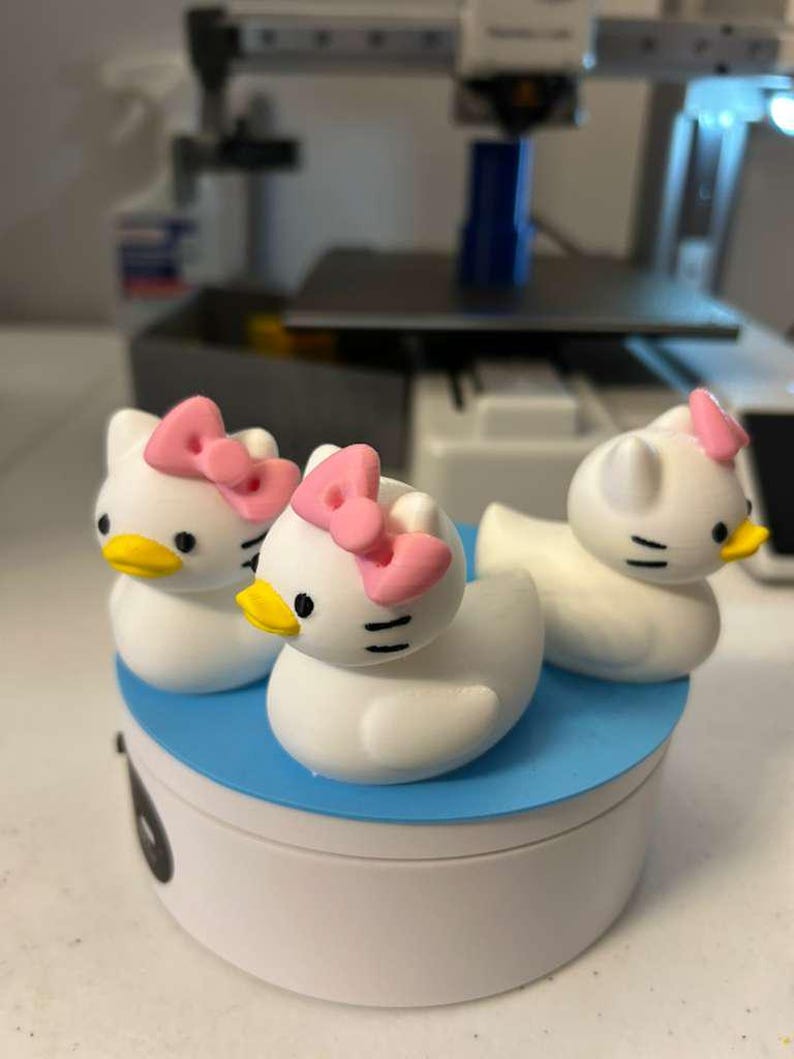 May include: Three white duck-shaped figurines with cat-like ears, black eyes, yellow beaks, and pink bows on their heads. The figurines are arranged on a circular blue and white platform, displaying a hybrid design of a duck and a cat character.