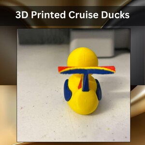 May include: A yellow 3D printed cruise duck figurine featuring a blue and red spoiler attached to its back. The text 3D Printed Cruise Ducks is displayed above the object. The duck has blue wing-like accents on its sides.