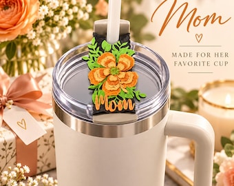 May include: A white tumbler with a clear lid and a white straw. Attached to the straw is a black topper featuring a layered orange flower, green leaves, and the word "Mom" in orange script. The text "Mom" and "MADE FOR HER FAVORITE CUP" appear nearby.