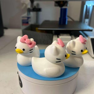 May include: Three white duck-shaped figurines with cat-like features, including pointed ears and whiskers. Each figurine has a small pink bow on its head and a yellow beak. The three figures are arranged on a round, white and blue platform.