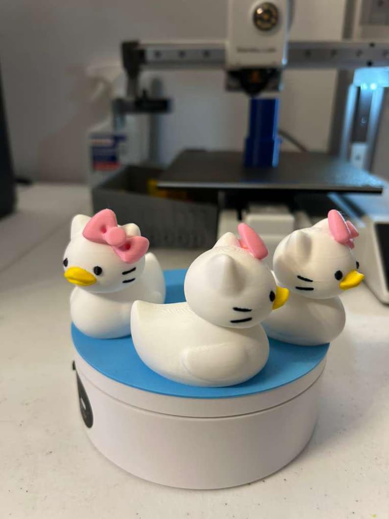 May include: Three white duck-shaped figurines with cat-like features, including pointed ears and whiskers. Each figurine has a small pink bow on its head and a yellow beak. The three figures are arranged on a round, white and blue platform.
