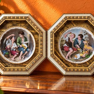 May include: Two octagonal wall plaques featuring circular porcelain plates with detailed paintings of children eating fruit. The frames are cream-coloured with dark wood-toned inner borders and decorative linear patterns.
