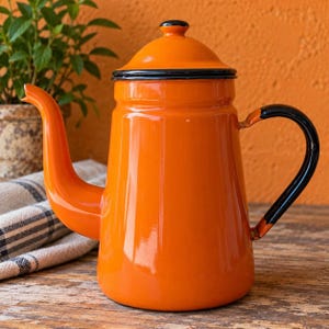 Puede incluir: Una cafetera de esmalte color naranja brillante con acabado lustroso, que presenta una tapa a juego con un pequeño pomo, un pico curvo y un asa negra. El borde de la tapa y la base del asa están acentuados con un fino detalle de borde negro.
