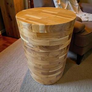 Puede incluir: Una mesa auxiliar de madera marrón claro con forma cilíndrica. La mesa está construida con pequeños trozos rectangulares de madera, creando una superficie texturizada. La parte superior es una pieza de madera circular y maciza. La mesa mide aproximadamente 51 cm de altura.