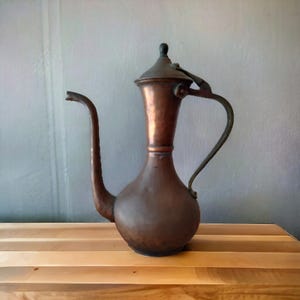 May include: A vintage copper pitcher with a long, curved spout and a handle. The pitcher has a bulbous base and a lid with a small, round knob. The copper has a rich, aged patina, suggesting it is an antique or a well-loved piece.