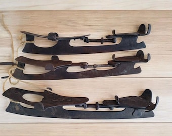 May include: Three pairs of antique ice skates with dark metal blades and adjustable bindings. The skates are arranged on a light wooden surface, with twine attached to some of them. The blades show signs of age and wear.