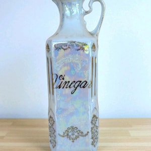 May include: Iridescent white ceramic vinegar bottle with a handle and the word "Vinegar" in black script. The bottle has gold accents and decorative floral patterns. A vintage kitchenware item.