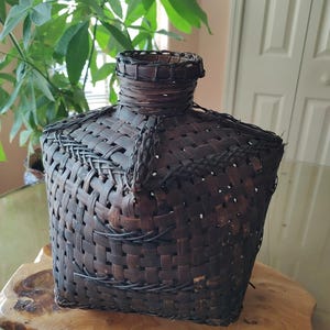 Könnte beinhalten: Ein quadratischer Korb aus dunkelbraunem geflochtenem Material mit einem geflochtenen Deckel. Der Korb ist aus natürlichen Materialien gefertigt und hat ein rustikales Aussehen.