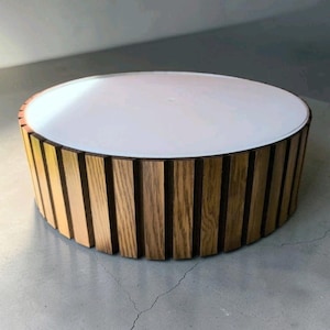 May include: A round, wooden table with a white top. The sides are made of vertical wooden slats. The table is a light brown color and sits on a gray concrete floor. The table is a modern design.