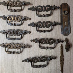 Op de afbeelding: Antieke bronzen lade-trekkers en hardware. De collectie bevat zeven sierlijke trekkers met een sleutelgatontwerp, een deurplaat met een sleutelgat, een kleine knop en een decoratieve handgreep. Gerangschikt op een licht houten oppervlak.