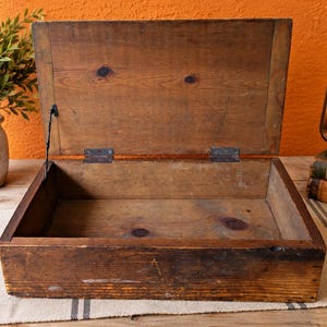 Op de afbeelding: Een rechthoekige houten doos met een opengeklapt deksel. De doos is gemaakt van natuurlijk bruin hout met zichtbare nerven, noesten en metalen scharnieren. Een dunne zwarte metalen ketting verbindt het deksel met de onderkant van de doos.