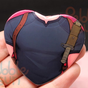 May include: Heart-shaped button featuring a stylized illustration of a dark blue shirt with brown straps and a tan knife. The button has a pink border and the text "ebble sun" is visible.