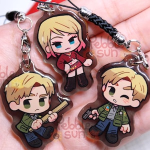 以下が含まれることがあります： 透明なアクリル製のキーホルダー3個。キャラクターは、ピンクの髪と赤い服を着た女性と、金髪の男性2人。女性は銃を持ち、一人の男性はバットを持ち、もう一人の男性は銃を持っている。キーホルダーは、銀色の留め具が付いた黒い紐で繋がっている。