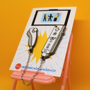 Puede incluir: Dos pines de esmalte en una tarjeta de presentación. Un pin es un mando a distancia de Wii gris y blanco, el otro es un nunchuk de Wii gris y blanco. Ambos pines están unidos a una cadena plateada. La tarjeta tiene un dibujo de dos figuras y un cubo de basura.