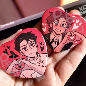 May include: Two heart-shaped pins with cartoon illustrations of two people making heart shapes with their hands. The pin on the left features a person with black hair and a white shirt. The pin on the right features a person with red hair and a black shirt.