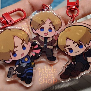 May include: Three clear acrylic keychains featuring a cartoon character with blonde hair. The character is wearing different outfits in each keychain. The keychains are attached to red metal key rings. The text "pebble sun shop" is printed on the bottom of one of the keychains.