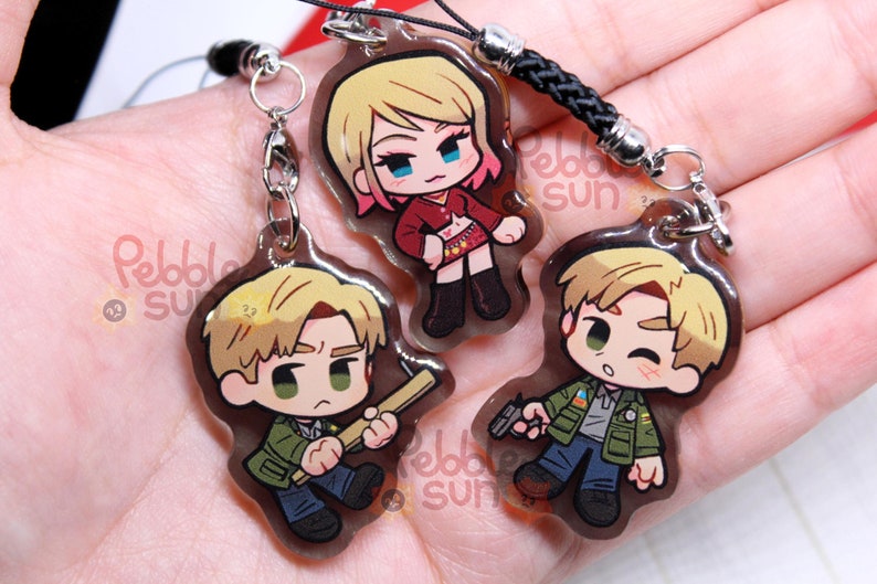May include: Three clear acrylic keychains featuring cartoon characters. The characters are a woman with pink hair and a red outfit, and two men with blonde hair. The woman is holding a gun, one man is holding a baseball bat, and the other man is holding a gun. The keychains are attached to black cords with silver clasps.