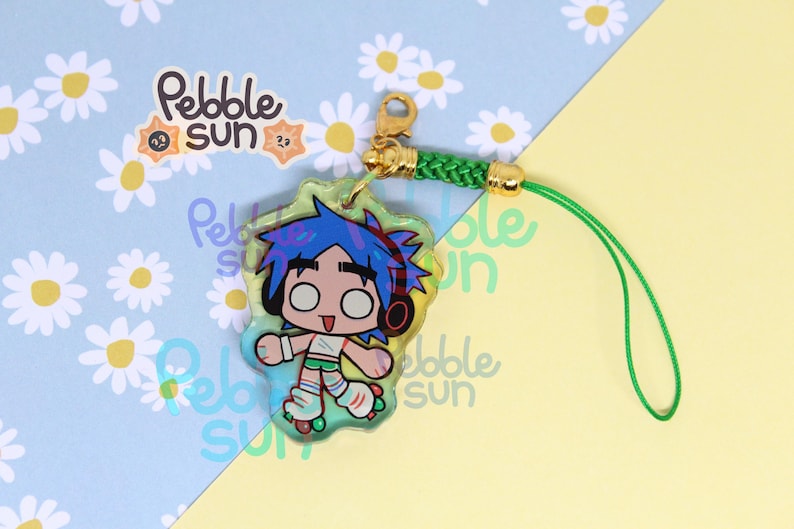 May include: A clear acrylic charm featuring a cartoon character with blue hair, headphones, and roller skates. The charm has a green border and is attached to a green cord with a gold clasp. The background has a daisy pattern and the text "Pebble Sun".