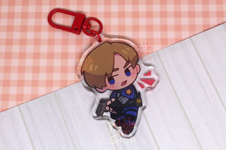 May include: A clear acrylic keychain featuring a cartoon character with brown hair, blue eyes, and a blue and black uniform. The character is holding a gun and has a red star on their shoulder. The keychain has a red metal ring.