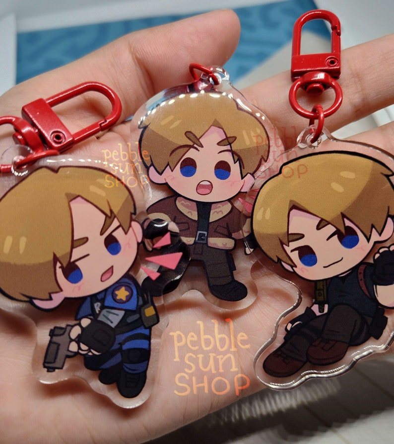 May include: Three clear acrylic keychains featuring a cartoon character with blonde hair. The character is wearing different outfits in each keychain. The keychains are attached to red metal key rings. The text "pebble sun shop" is printed on the bottom of one of the keychains.