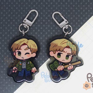 May include: Two cartoon character keychains with silver key rings. The characters are male with brown hair and are wearing different outfits. One character is holding a gun and the other is holding a knife.