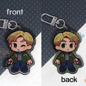 May include: A keychain featuring a cartoon character with brown hair, wearing a green jacket and blue jeans. The keychain is made of clear acrylic and has a silver metal ring.
