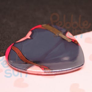 May include: Heart-shaped resin badge featuring a stylized illustration. The design includes a navy blue element with a brown strap and a light brown object. The badge has a pink border and is set against a pink surface with heart patterns.