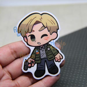 May include: A chibi-style sticker featuring a cartoon character with blonde hair, a green jacket, and a gray shirt, holding a gun. The character has a small cut on their cheek. The sticker is held in a hand.