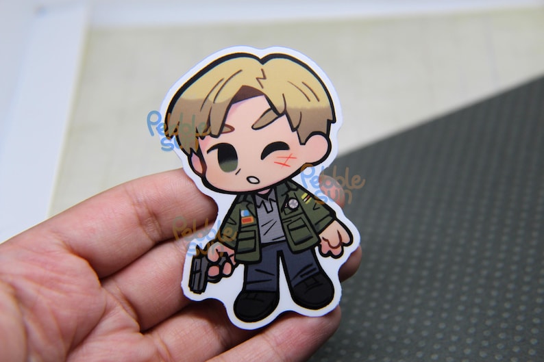 May include: A chibi-style sticker featuring a cartoon character with blonde hair, a green jacket, and a gray shirt, holding a gun. The character has a small cut on their cheek. The sticker is held in a hand.