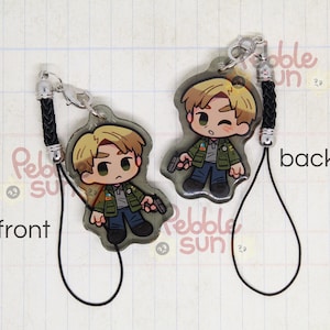 May include: A black and white illustrated character keychain with a black cord. The character is a young man with blond hair, wearing a green jacket and holding a gun. The keychain is labeled "front" and "back".