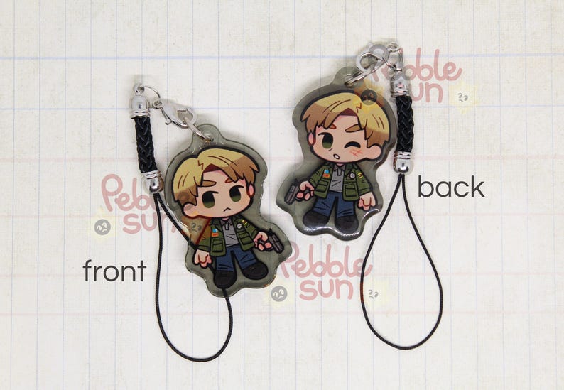 May include: A black and white illustrated character keychain with a black cord. The character is a young man with blond hair, wearing a green jacket and holding a gun. The keychain is labeled "front" and "back".