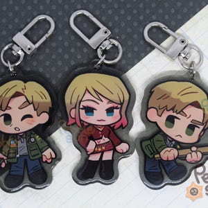 May include: Three cartoon character keychains with silver key rings. The characters are a woman with pink hair wearing a red top and a black skirt, a man with brown hair wearing a green jacket, and a man with brown hair wearing a green jacket and holding a wooden stick.