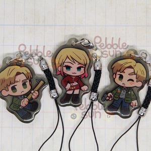 May include: Three character charms with black cords. The charms are round and feature cartoon characters. One charm features a blonde woman with a red top and black skirt. Another charm features a man with brown hair and a green jacket. The third charm features a man with brown hair and a blue jacket.