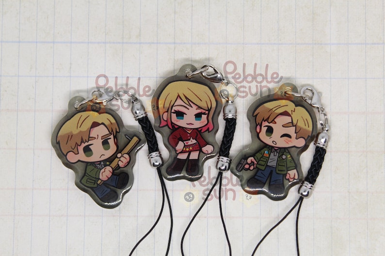 May include: Three character charms with black cords. The charms are round and feature cartoon characters. One charm features a blonde woman with a red top and black skirt. Another charm features a man with brown hair and a green jacket. The third charm features a man with brown hair and a blue jacket.