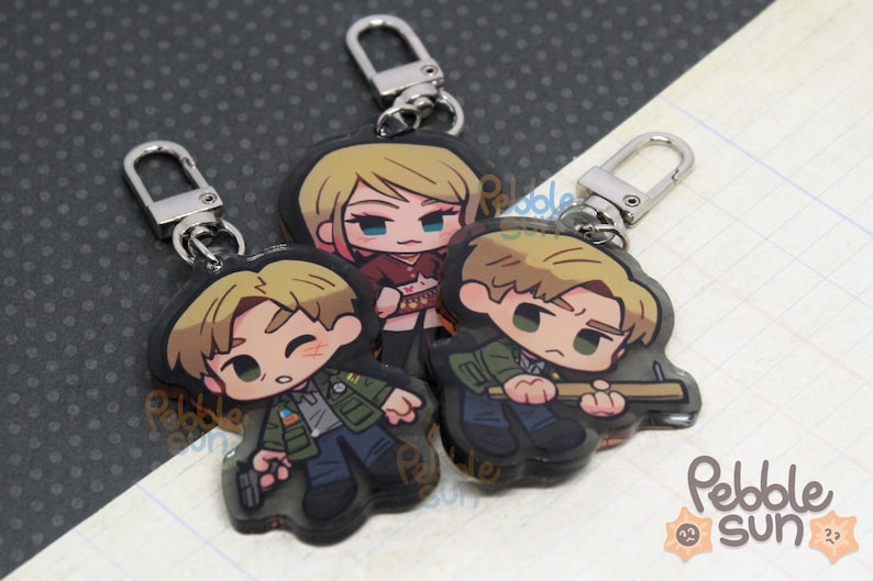 May include: Three character keychains featuring cartoon illustrations of three people. The characters are a woman with blonde hair wearing a red and black outfit, a man with blonde hair wearing a green jacket and blue jeans, and a man with brown hair wearing a green jacket and blue jeans. The keychains are made of clear acrylic and have silver metal key rings.