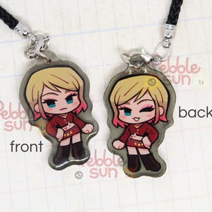 May include: A double-sided charm featuring a cartoon character with blonde hair, pink highlights, and a red and white outfit. The charm is attached to a black cord and has the words "Pebble Sun" on the back.