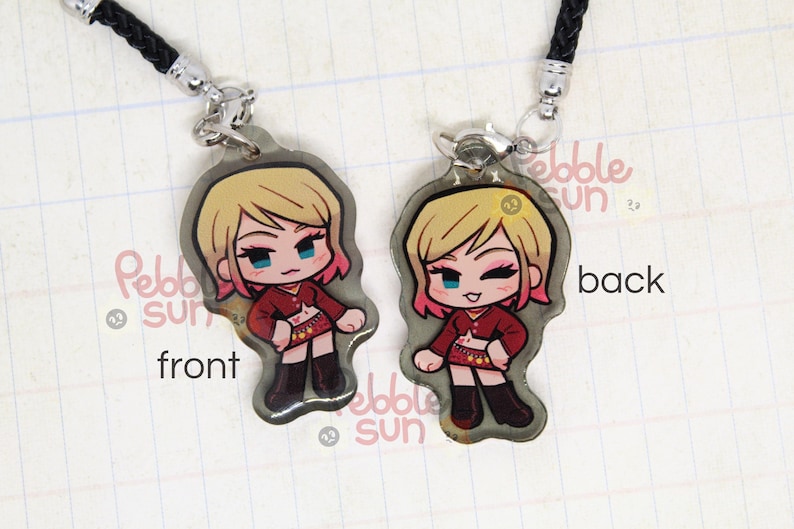 May include: A double-sided charm featuring a cartoon character with blonde hair, pink highlights, and a red and white outfit. The charm is attached to a black cord and has the words "Pebble Sun" on the back.
