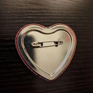 May include: A heart-shaped pinback button with a safety pin on the back. The button has a metallic silver center and a red and pink border. The button is on a dark wood surface.