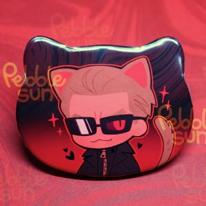 May include: A cat-shaped button featuring a cartoon illustration of a person with sunglasses and a black jacket. The background is red with the text "Pebble Sun". The character has one red eye and one black eye.