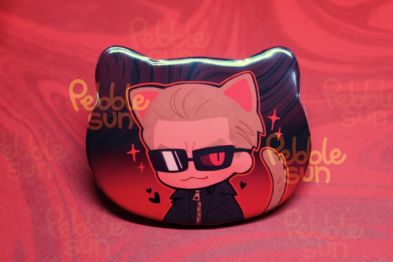 May include: A cat-shaped button featuring a cartoon illustration of a person with sunglasses and a black jacket. The background is red with the text "Pebble Sun". The character has one red eye and one black eye.