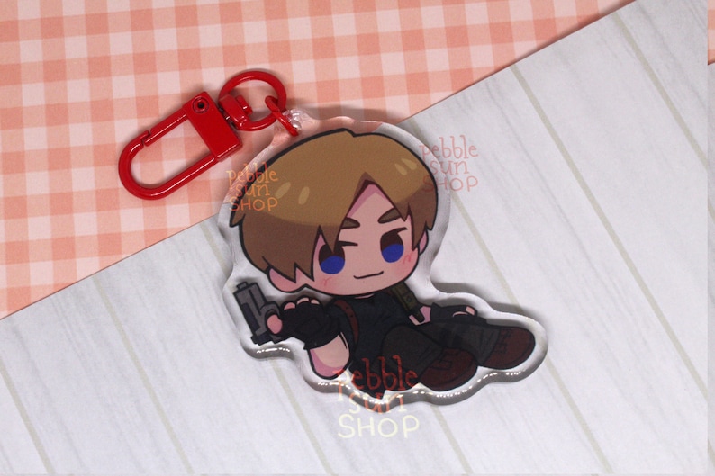 May include: A clear acrylic keychain featuring a cartoon character holding a gun. The character has brown hair, blue eyes, and is wearing a black shirt and pants. The keychain is attached to a red metal ring.