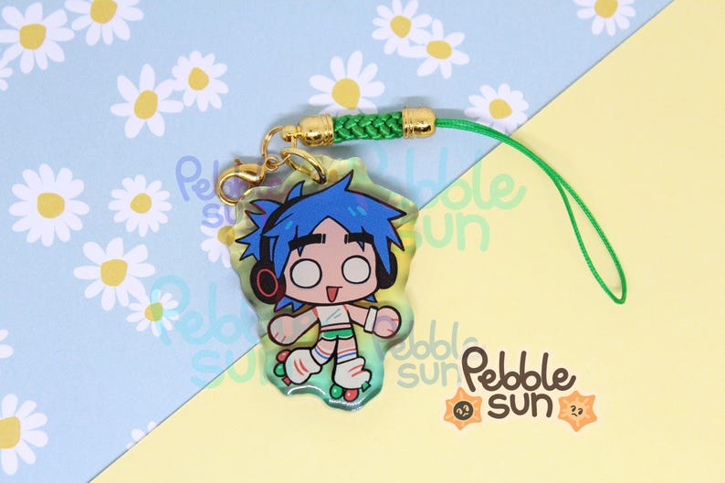May include: A clear acrylic charm featuring a cartoon character with blue hair, headphones, and roller skates. The charm has a gold clasp, green cord, and a green and gold braided accent. The background is light blue with white daisy patterns and yellow.
