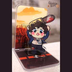 May include: A clear acrylic stand featuring a cartoon character holding a chainsaw with blood splatter. The character is wearing a blue suit and has a surprised expression. The background depicts a sunset scene with trees and the text "Pebble Sun".