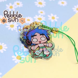 May include: A clear acrylic charm featuring a cartoon character with blue hair, headphones, and roller skates. The charm has a green lanyard and gold-colored hardware. The charm is from Pebble Sun.