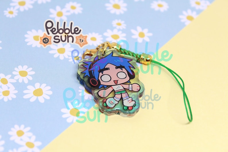 May include: A clear acrylic charm featuring a cartoon character with blue hair, headphones, and roller skates. The charm has a green lanyard and gold-colored hardware. The charm is from Pebble Sun.