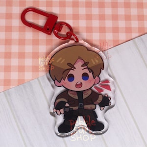 May include: A clear acrylic keychain featuring a cartoon character with brown hair, a blue jacket, and a red scarf. The character is holding a gun and has a surprised expression on their face. The keychain has a red metal ring.